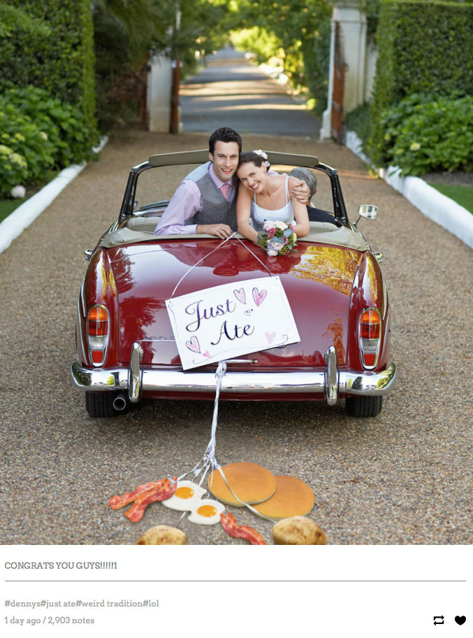 A couple, presumably husband and wife, embrace in the back of a car with a sign hanging on the back that says, "Just Ate." Instead of cans being dragged behind the vehicle, several breakfast foods are - including bacon, eggs, and pancakes