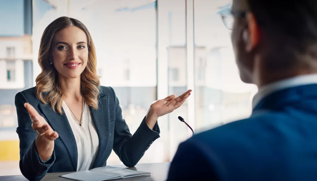 A woman in business attire sitting at a table across from a man in a suit, in an interview situation